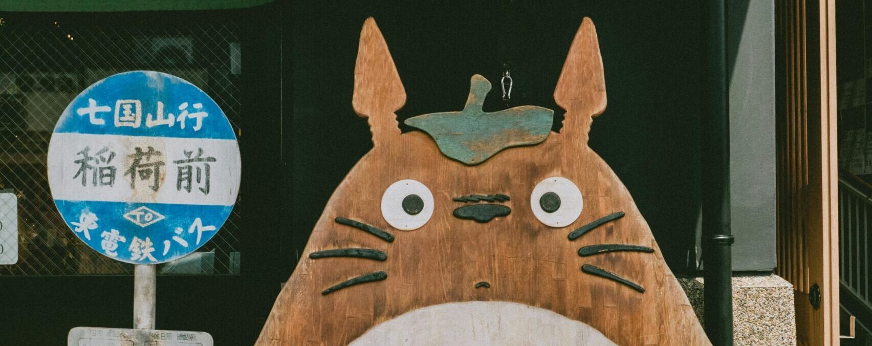 Wooden Totoro sculpture on a sunny Japanese street, next to local signage.